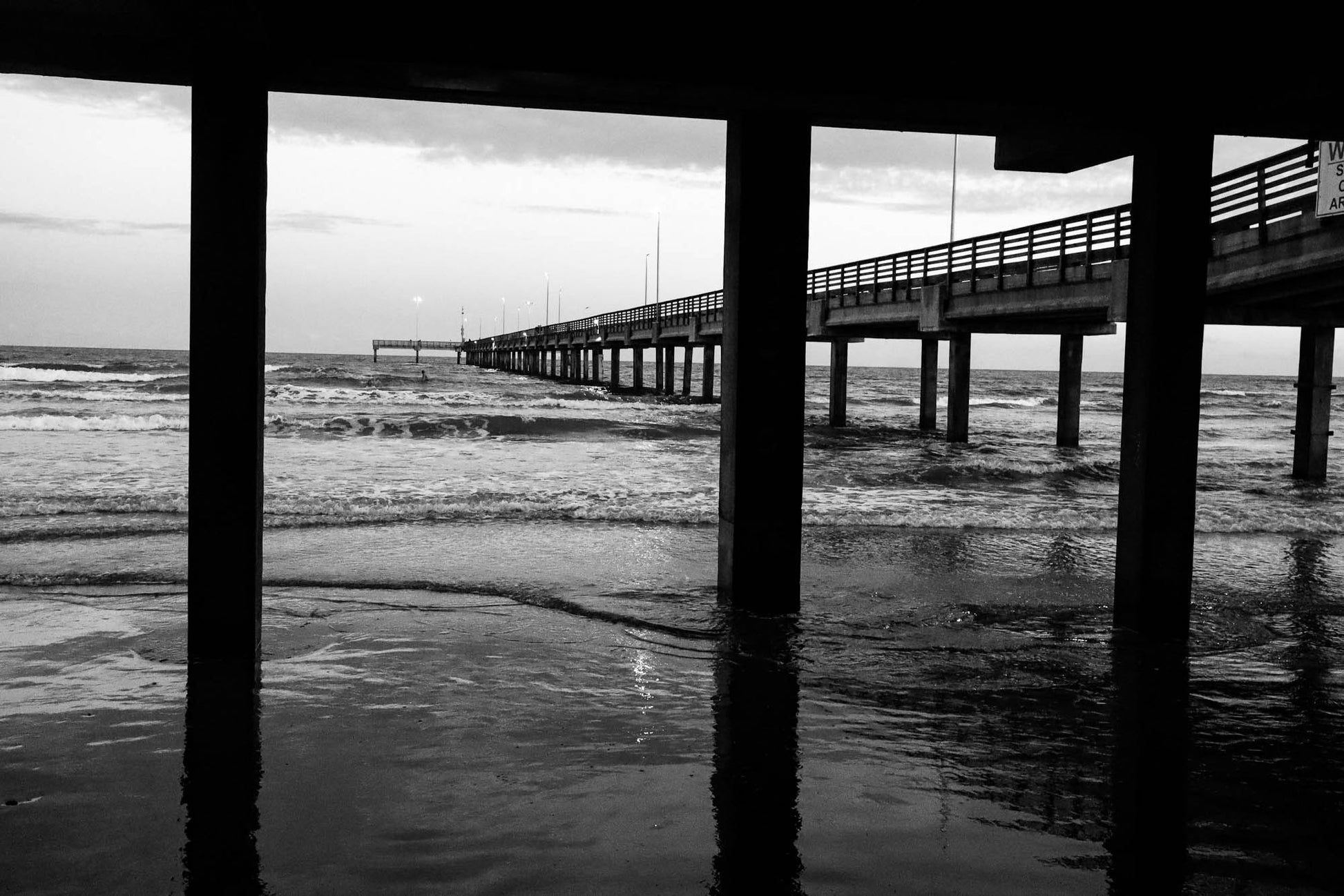 Black and white photo of a pier extending into the ocean from under a wooden structure.