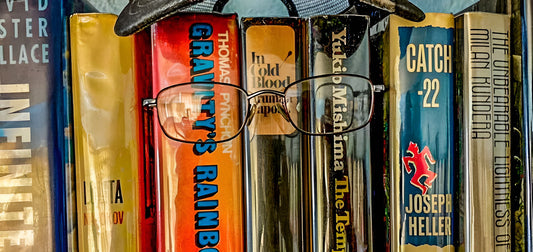 Stack of books with a pair of glasses on top, featuring titles like 'Gravity's Rainbow' and 'Catch-22'.