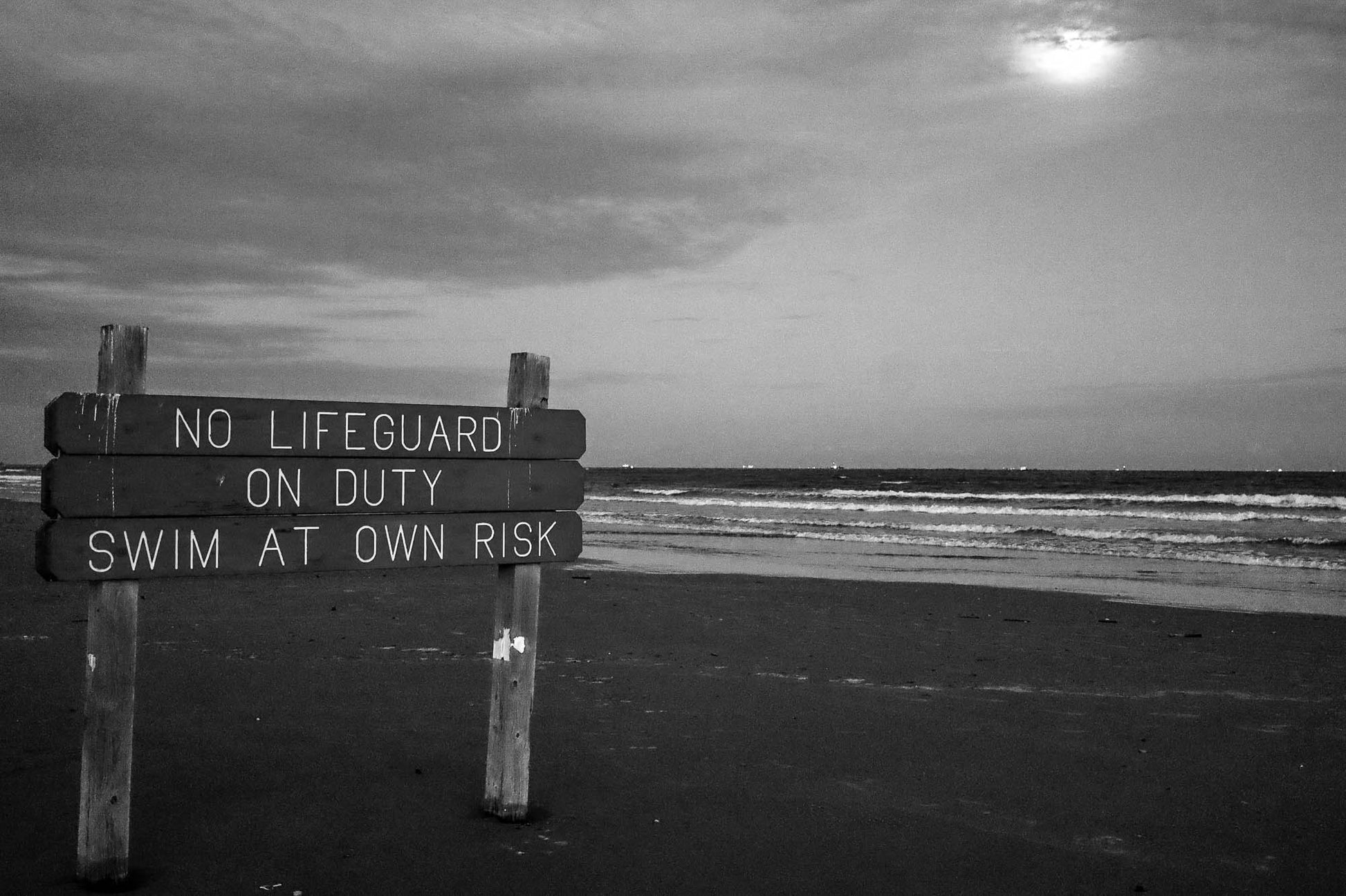 Wooden sign on a beach warning of no lifeguard on duty and swim at your own risk, with ocean and cloudy sky in the background.