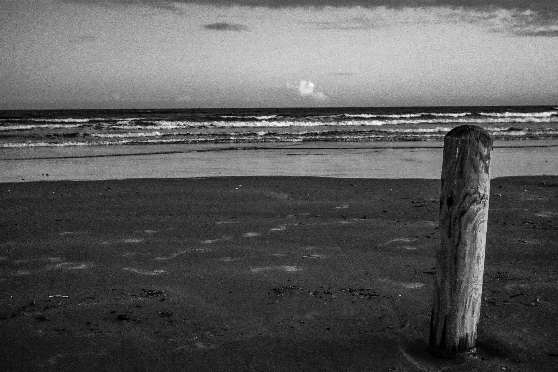 Black and white photo of a beach with a wooden post and ocean waves.