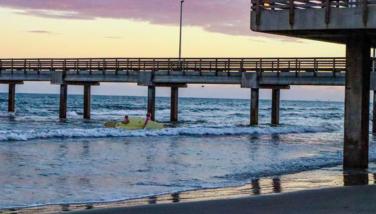 Beach scene with a pier and surfers in the water at sunset.