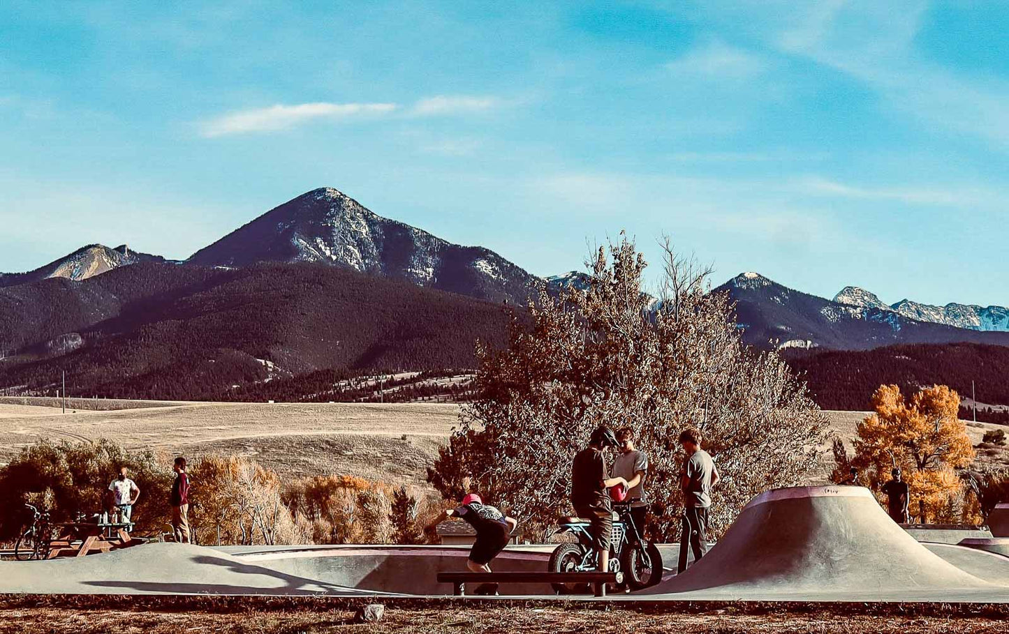 People with skateboards and bicycles in a skate park with mountains in the background