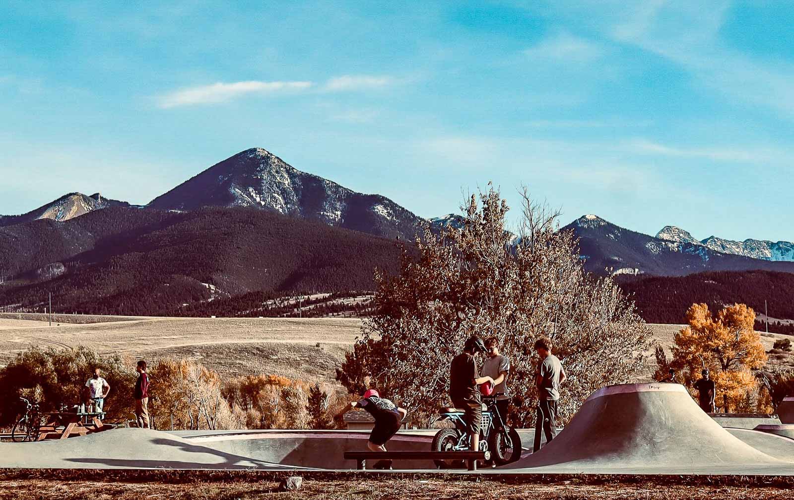 People with skateboards and bicycles in a skate park with mountains in the background
