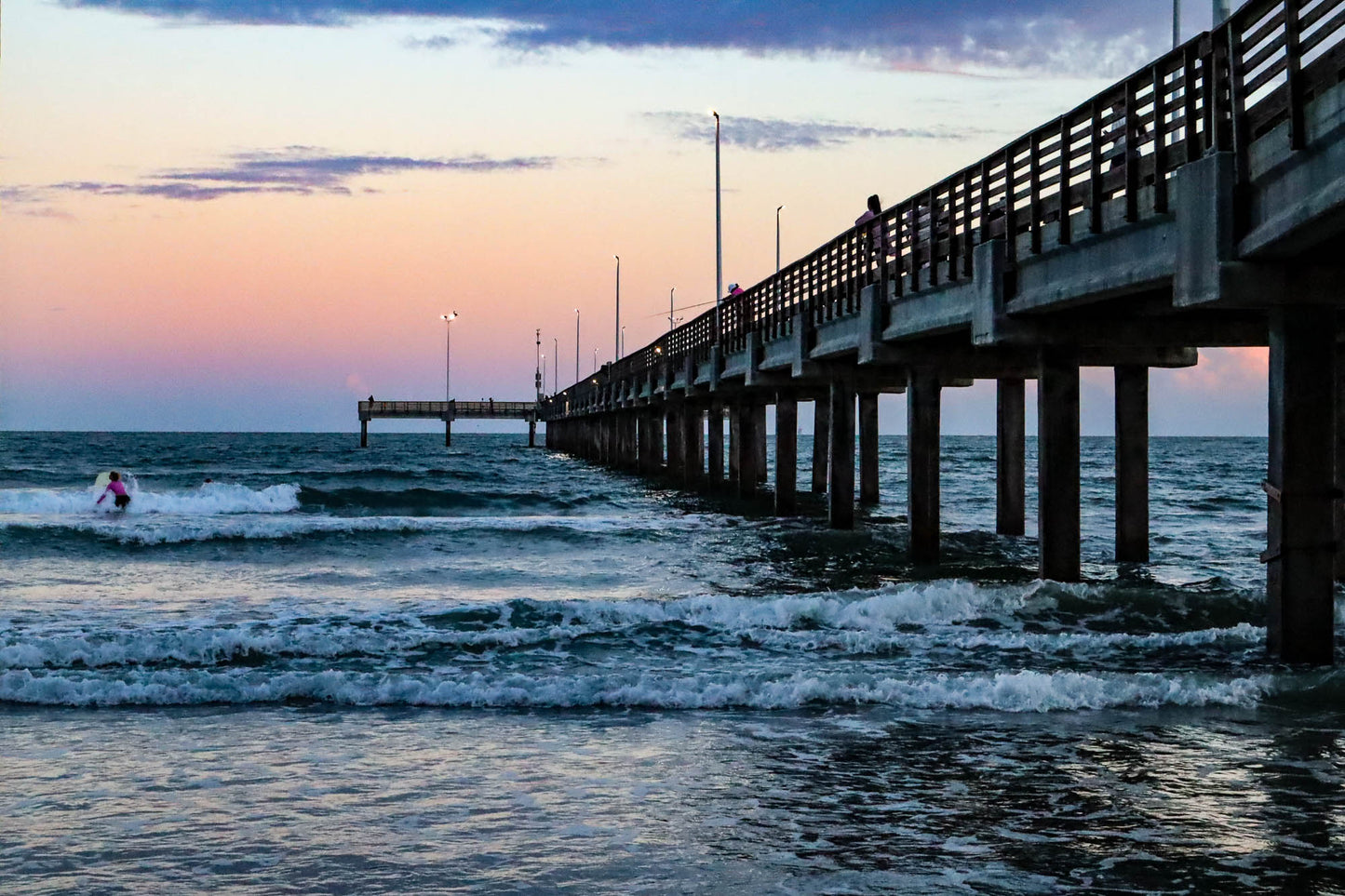 Pier extending into the ocean with a surfer in the water at sunset.