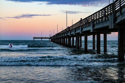 Pier extending into the ocean with a surfer in the water at sunset.